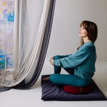 Kobieta siedzi po turecku na malinowej poduszce do medytacji na fioletowej macie; w tle kolorowy obraz oparty o ścianę i opadające zasłony.